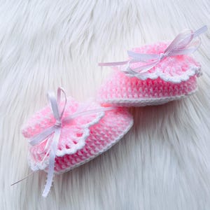 May include: A pair of handmade baby booties. The booties are pink with a white scalloped trim and white ribbon ties. The booties are made of a soft yarn and are lying on a white, fluffy surface.