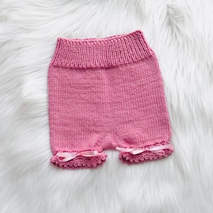 Puede incluir: Unos pantalones cortos de punto de color rosa con una cintura acanalada y un dobladillo texturizado en cada abertura de las piernas. Cada pierna está decorada con un pequeño lazo de cinta de satén rosa claro atado en el borde festoneado.