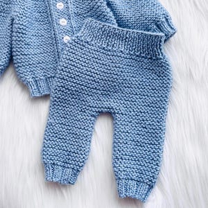 May include: A light blue knitted baby cardigan and matching pants set. The cardigan has a button closure and the pants have a simple design.