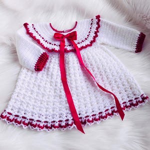 May include: A white crochet baby dress with red trim and a red satin bow. The dress has long sleeves and a scalloped collar. The dress is displayed on a white, fluffy surface.