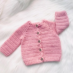 以下が含まれることがあります： 手作りの、明るいピンク色のベビー用かぎ針編みカーディガン。長袖で、丸いネックラインがあり、5つの星型の装飾ボタンで留められています。白いふわふわした表面に置かれています。