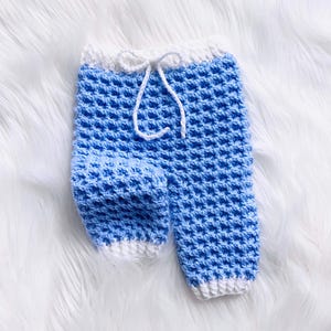 Op de afbeelding: Handgemaakte babybroek in een lichtblauw wafelpatroon met witte afwerking. De broek heeft een taille met trekkoord en wordt gepresenteerd op een witte, pluizige ondergrond. Een charmant en gezellig item voor een pasgeborene.