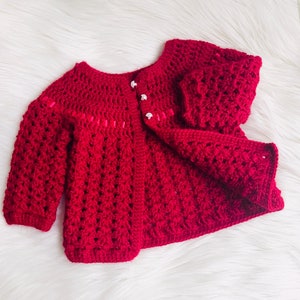 May include: A red crocheted baby cardigan with white buttons and pink trim. The cardigan is laid out on a white fluffy surface.