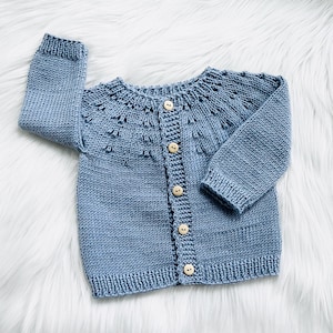 Könnte beinhalten: Hellblau gestrickte Baby-Strickjacke mit rundem Ausschnitt und langen Ärmeln. Die Strickjacke hat eine Knopfleiste mit Holzknöpfen und dekorativen Nähten am Halsausschnitt. Das Kleidungsstück ist aus weichem Garn gefertigt.