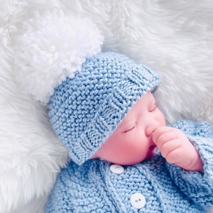 May include: A baby wearing a light blue knitted hat with a white pom-pom and a matching light blue knitted cardigan.