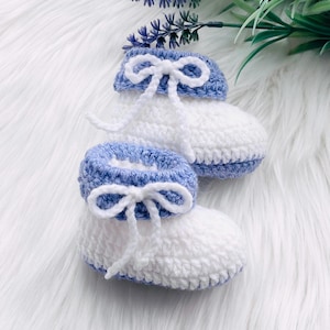 May include: A pair of crocheted baby booties in white and light blue with white bows. The booties are on a white fluffy surface.