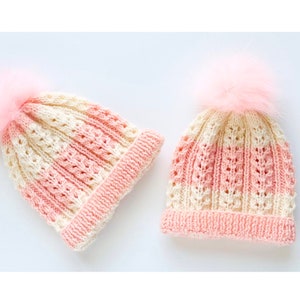 May include: Two knitted hats with pink and cream stripes and fluffy pom poms. The hats are made with a lacy stitch pattern.
