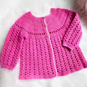 May include: A bright pink, handmade crochet cardigan with long sleeves and a round neckline. The openwork design features a scalloped yoke and is fastened with three wooden buttons. The cardigan is displayed on a white, fluffy surface.