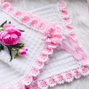 May include: A white crocheted baby blanket with a pink scalloped border and accents. A pink ribbon with a decorative embellishment is attached. A pink peony and bud are placed near the blanket.