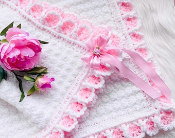 May include: A white crocheted baby blanket with a pink scalloped border and accents. A pink ribbon with a decorative embellishment is attached. A pink peony and bud are placed near the blanket.