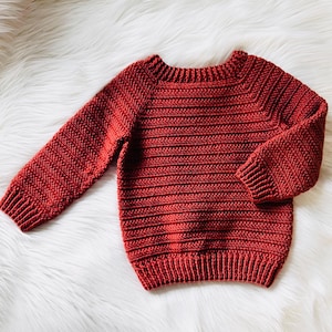 Może przedstawiać: Czerwony dzianinowy sweter z długimi rękawami. Sweter ma prążkowany dół i mankiety oraz okrągły dekolt. Wzór dzianiny jest poziomy na ciele i pionowy na rękawach. Sweter jest wykonany z miękkiej, teksturowanej przędzy.