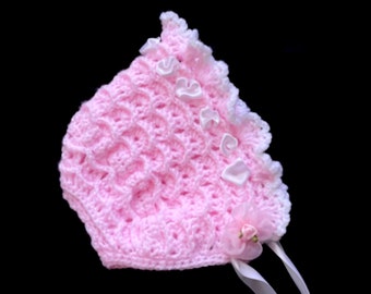 May include: A pink crocheted baby bonnet with white lace trim and white satin ribbon ties. The bonnet has a flower embellishment.