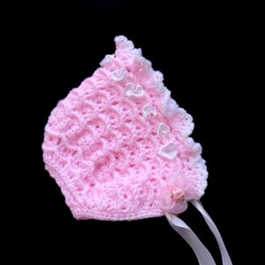 May include: A pink crocheted baby bonnet with white lace trim and white satin ribbon ties. The bonnet has a flower embellishment.