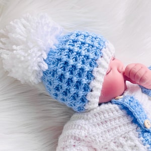 May include: A blue and white crocheted baby hat with a white pom-pom on top. The hat is worn by a baby doll with a blue and white crocheted cardigan.