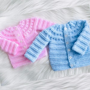 May include: Two knitted baby cardigans, one pink and one light blue, with button fastenings. The cardigans have a striped pattern and are laid out on a white fluffy surface.