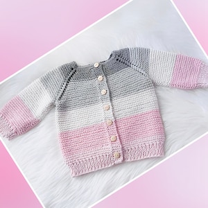 Puede incluir: Un cárdigan de punto a rayas para bebé con cierre de botones. El cárdigan es rosa, blanco y gris.