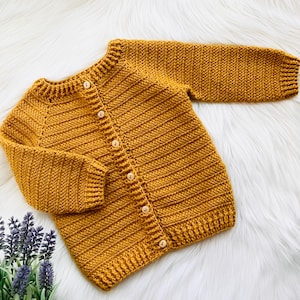 May include: Mustard yellow knitted cardigan with long sleeves and wooden buttons. The handmade sweater is displayed on a white fluffy surface. The cardigan is a cozy, warm garment for children.