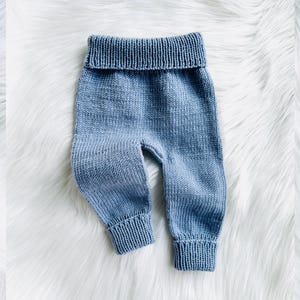 Pode incluir: Calças de bebê de malha azul claro com cós dobrado e punhos canelados. As calças são feitas de um fio macio e texturizado. As calças de bebê são exibidas em uma superfície branca e fofa.