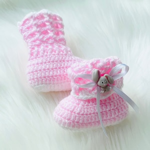 May include: A pair of pink crocheted baby booties with a white trim. One bootie has a white ribbon and a gray bunny button.