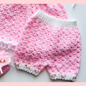 May include: A pink crocheted baby outfit with a white trim and pink flower embellishments. The outfit includes a hat, shorts, and booties.