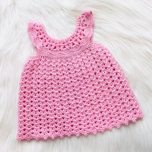 May include: A pink crocheted baby dress with a delicate lace pattern. The dress has a sleeveless design and a scalloped hem.