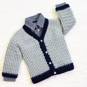 May include: A gray and navy blue crocheted cardigan with a button closure. The cardigan has a V-neck and a textured stitch pattern.
