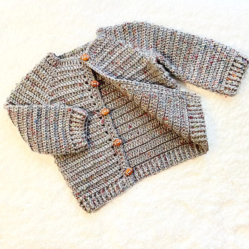May include: A gray and white crocheted cardigan with brown football-shaped buttons. The cardigan has a speckled pattern and is made with a soft yarn.