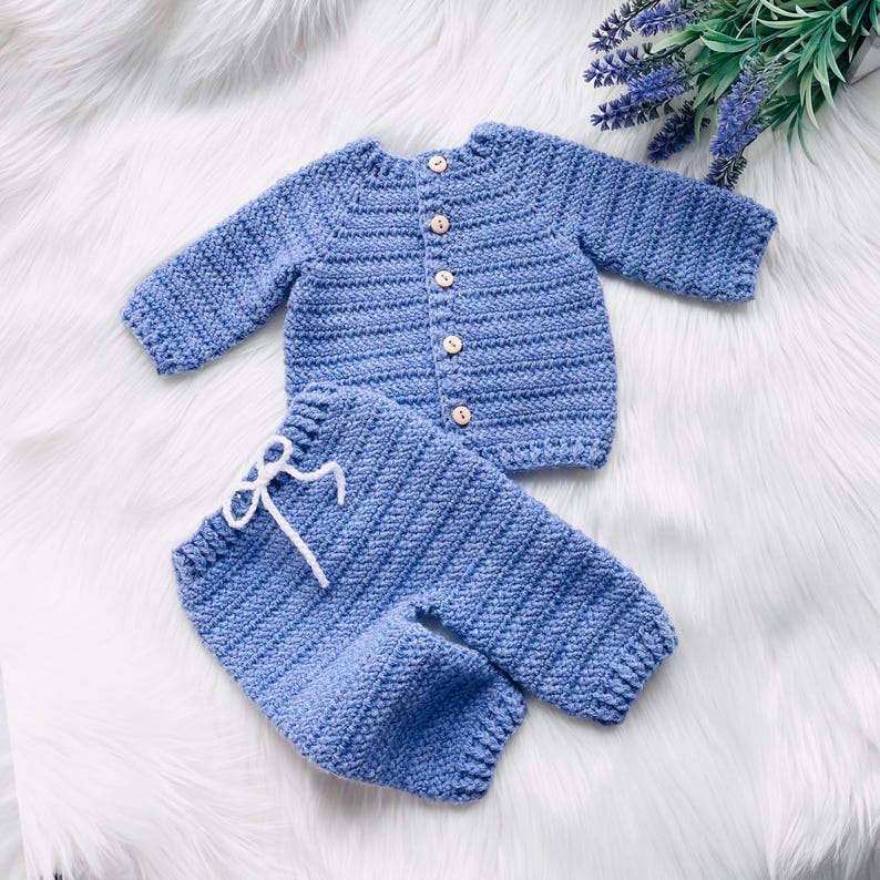 May include: A blue crocheted baby outfit with a cardigan and pants. The cardigan has a button closure and the pants have a white ribbon tie.