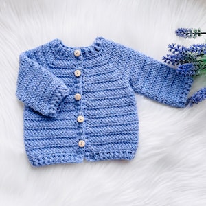 May include: A blue knitted baby cardigan with button closures. The cardigan is made with a soft, textured yarn.