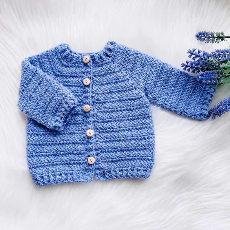 May include: A blue knitted baby cardigan with button closures. The cardigan is made with a soft, textured yarn.