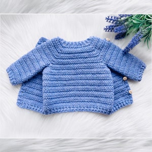May include: A blue crocheted baby cardigan with button closures. The cardigan is laid flat on a white fuzzy surface.