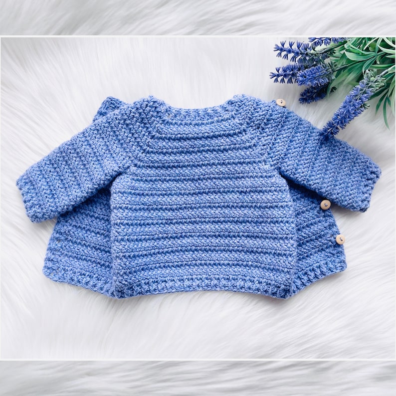 May include: A blue crocheted baby cardigan with button closures. The cardigan is laid flat on a white fuzzy surface.