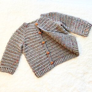 May include: A gray crocheted cardigan with brown buttons. The cardigan has a speckled pattern and is made with a soft yarn.
