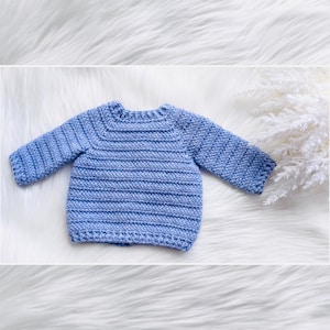 May include: A light blue crocheted baby sweater with long sleeves. The sweater is laid flat on a white fluffy surface.