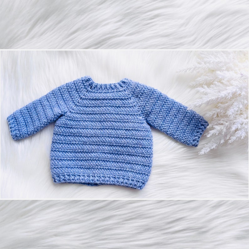 May include: A light blue crocheted baby sweater with long sleeves. The sweater is laid flat on a white fluffy surface.