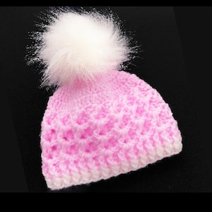 May include: A pink crocheted beanie with a white pom pom. The beanie has a textured stitch pattern.