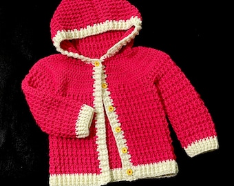 May include: A pink crocheted hooded cardigan with white trim and yellow buttons.