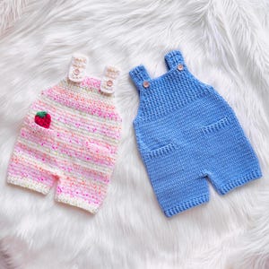 May include: Two handmade crochet baby overalls. One is pink and white with a red strawberry applique on the pocket. The other is solid blue. Both have button closures on the straps and pockets.