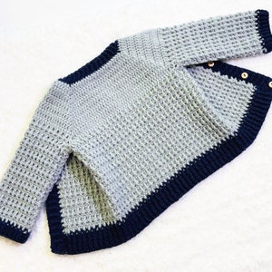 May include: A gray crocheted sweater with a navy blue trim. The sweater has a slightly asymmetrical hemline and is made with a textured stitch.