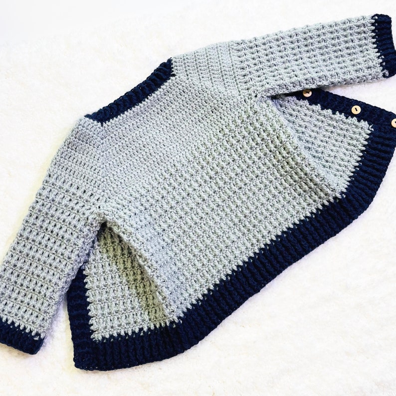 May include: A gray crocheted sweater with a navy blue trim. The sweater has a slightly asymmetrical hemline and is made with a textured stitch.