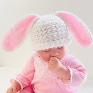 May include: A white crocheted bunny hat with pink ears. The hat is on a baby's head.