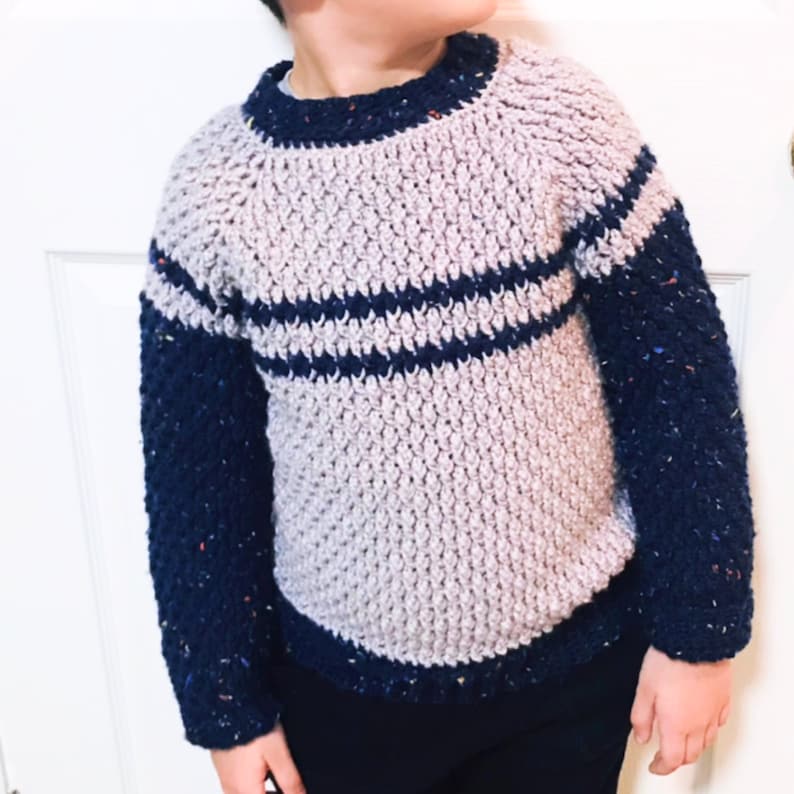 May include: A crocheted sweater with a light purple body and navy blue sleeves. The sweater has two horizontal stripes of navy blue across the chest.