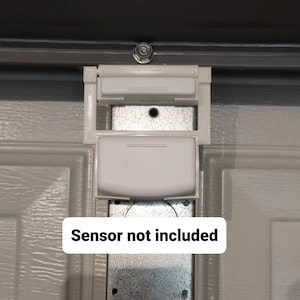 May include: White garage door sensor mount with a metal bracket and a bolt. The text "Sensor not included" is displayed on a white rectangle at the bottom of the image. The mount is attached to a gray garage door.