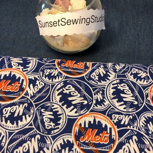May include: A blue and white fabric with a repeating pattern of the New York Mets baseball team logo. The logo includes the word "Mets" in orange and white, with a baseball and a skyline design. The text "SunsetSewingStudio" is also visible.