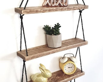 May include: Three wooden shelves hanging from black ropes, with a small succulent plant in a gray pot on the middle shelf, a yellow alarm clock on the bottom shelf, and a gold 'XOXO' on the top shelf.