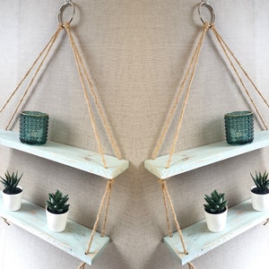 May include: Two-tier hanging shelf made of light blue painted wood, suspended by natural rope and a silver ring. The top shelf holds a teal glass candle holder. The bottom shelf displays two small potted succulents.