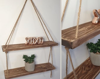 May include: Two wooden shelves hanging from the ceiling with rope. The shelves are stained a dark brown and have a rustic look. The top shelf has a small gold 'XOXO' sign on it. The bottom shelf has a small green plant in a gray pot.