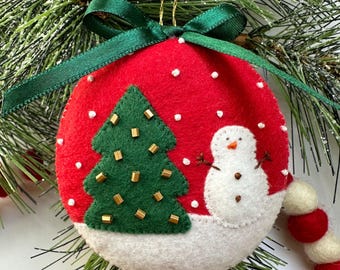 May include: Red felt Christmas ornament with a green Christmas tree and a snowman. The tree is decorated with gold beads. The ornament has a white snow base and is topped with a green ribbon.
