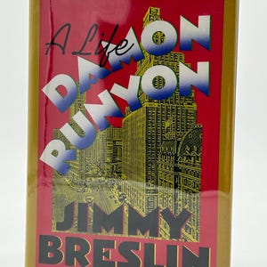 May include: A hardback book titled "A Life of Damon Runyon" by Jimmy Breslin. The cover has a red background with white and blue text, and a gold illustration of a city skyline. The book has a gold border.