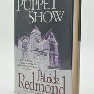 May include: A hardcover book titled "The Puppet Show" by Patrick Redmond. The cover features a dark background with a purple-toned illustration of a building. The author's name is in a burgundy color, and the book's title is in white.
